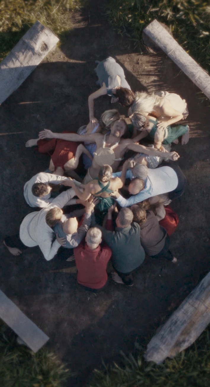 An overhead view of Wolf with arms extended, surrounded by a group inside four benches in a grassy clearing.